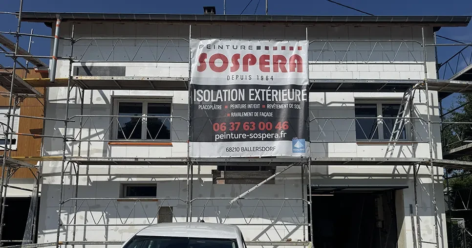 Isolation extérieure d’une maison à Strueth : façade avant avec échafaudage et banderole Peinture Sospera, entreprise RGE du Sundgau spécialisée en isolation de façade.